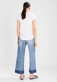 Bílý přiléhavý tričko a široké světle modré džíny s tmavomodrým lemem. Džíny mají třepený okraj a jsou spárované s vzorovanými nazouvacími botami.