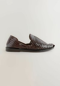 Zapatos marrones de cuero tipo slip-on con un diseño trenzado, punta redonda y suela plana. La textura es suave con una silueta estructurada.