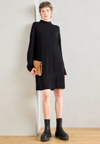 Melna adīta turtlenecks kleita ar garām piedurknēm, vidus augšstilba garumā. Saskaņota ar masīvām melnām potīšu zābakām un brūnu somiņu. Minimālistisks dizains.