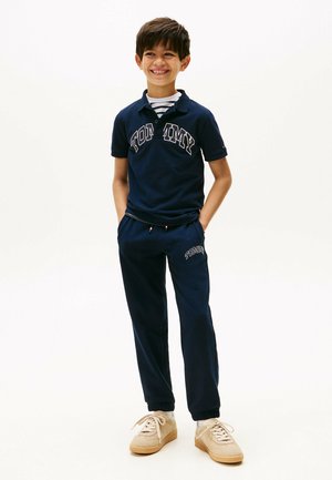 Polo bleu marine avec le logo "TOMMY", sous-couche rayée, pantalons de survêtement bleus assortis, poignets élastiques et baskets en daim beige.