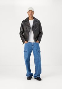 Svart läderbikerjacka med silverblixtlås, vit T-shirt, lösa blå denimcargo-byxor och svarta skor. Ljus keps på huvudet.