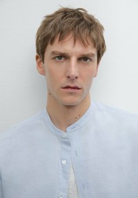 Camicia in lino azzurro chiaro con colletto a band, caratterizzata da una chiusura con bottoni davanti e una trama sottile, indossata sopra una t-shirt bianca.