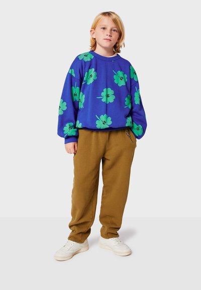 Felpa blu con motivi di trifoglio verdi, vestibilità ampia, abbinata a pantaloni della tuta marroni. Bambino che indossa sneaker bianche, in piedi su uno sfondo semplice.