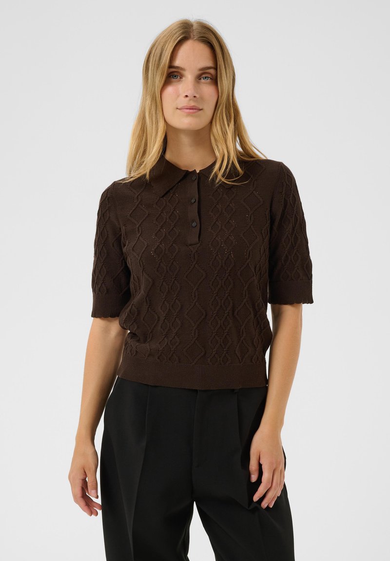 Jeune femme aux longs cheveux blonds portant un polo en maille à manches courtes marron foncé et un pantalon noir taille haute sur un fond uni.