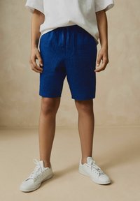 Shorts rayés bleus en tissu texturé, dotés d'une taille élastique et d'un cordon de serrage. Associés à des baskets blanches.
