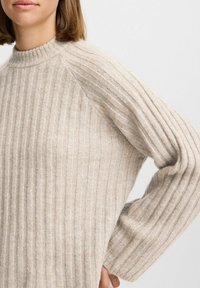 Beige ribbad tröja med hög krage och raglanärmar, som har en mjuk textur och en avslappnad passform. Texturdetaljerna är framträdande.
