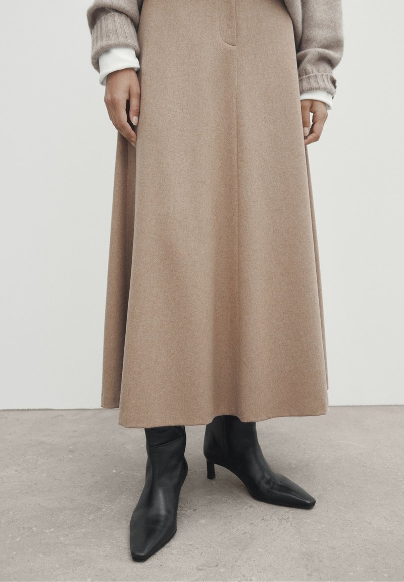 Personne portant une longue jupe beige, un pull beige, des poignets de chemise blancs et des bottines à talons noirs à bout pointu, debout sur un sol en béton.