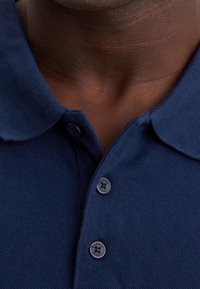 Marinblå pikétröja med en texturerad tyg, utrustad med krage och tre knappar med ett cirkulärt mönster på knäppningen.