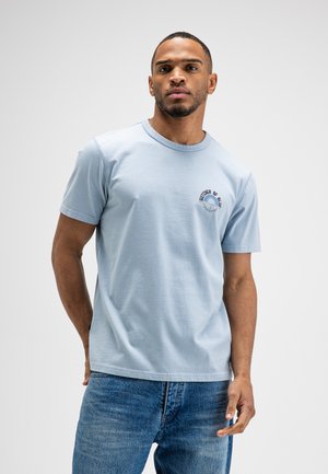 Man draagt een lichtblauw T-shirt met korte mouwen met een cirkelvormig tekstlogo op de borst en een blauwe jeans, staand tegen een effen achtergrond.