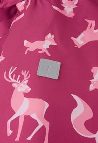 Vaaleanpunainen kangas, jossa on leikkisiä kuvastoja hirvistä ja supikoirista eri asennoissa, ja siinä on neliön muotoinen harmaa etiketti ommeltuna.