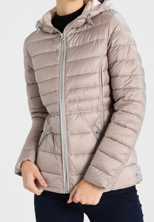 Person iført en lys beige kviltet pufferjakke med frontlynlås og sidelommer, kombineret med mørke bukser.