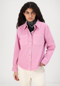 Pink button-up shirt with two chest pockets, tailored fit, and rounded hem. Worn over a black patterned bandana and a dark shirt.