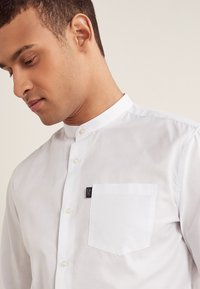 Hvid skjorte med knapper og mandarin-krave, brystlomme foran og sort logoetiket. Stoffet ser glat og skræddersyet ud.