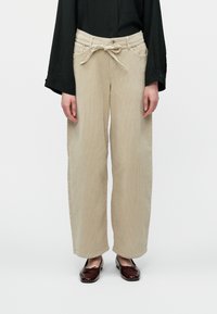 Moteris vilki kreminės spalvos odekolono kelnes su surištu diržu, juodą ilgomis rankovėmis viršutinę dalį ir tamsiai raudonus blizgius baleto batelius.