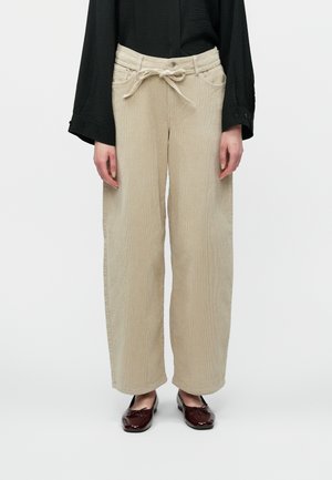 Egy nő bézs kordnadrágot visel, megkötős derékszíjjal, fekete hosszúujjú felsővel és sötétvörös lakkbőr ballerina cipővel.