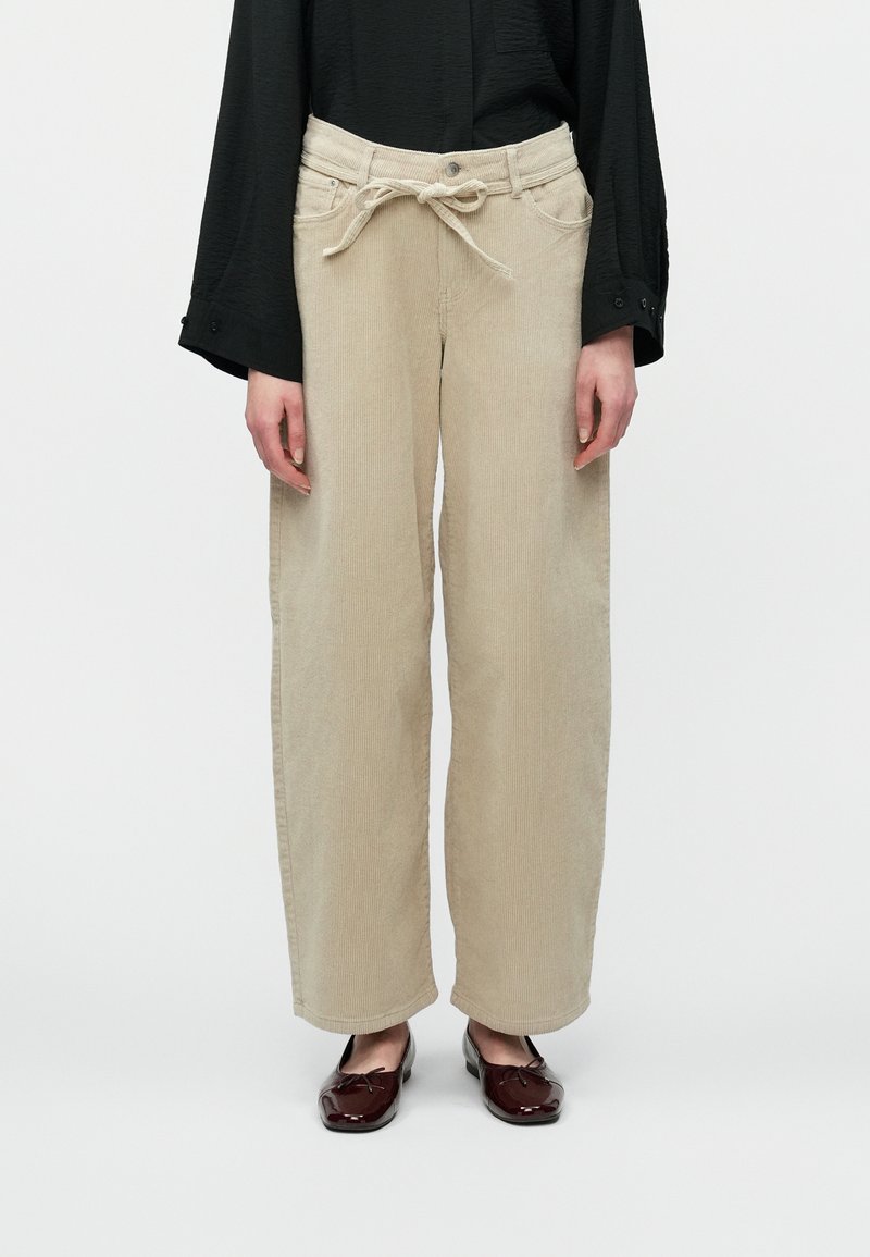 Moteris vilki kreminės spalvos odekolono kelnes su surištu diržu, juodą ilgomis rankovėmis viršutinę dalį ir tamsiai raudonus blizgius baleto batelius.