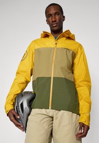 Wasserabweisende Jacke in Gelb, Olivgrün und Beige. Verfügt über einen Reißverschluss vorne, eine Kapuze und elastische Bündchen. Ein Fahrradhelm wird in einer Hand gehalten.