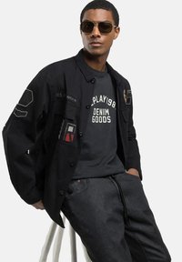 Chaqueta negra con parches, camiseta gris con texto blanco y pantalones de mezclilla oscuros. Modelo usando gafas de sol, de pie junto a un taburete blanco.