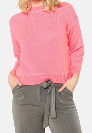 Personne portant un pull en laine rose vif et un pantalon gris taille haute noué avec une ceinture en tissu, mains dans les poches.