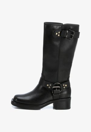 Bota de cuero negra hasta la pantorrilla con tacón bajo y cuadrado, correas con hebilla en la parte superior y en el tobillo, y detalles de remaches metálicos.