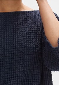 Mörkblå, strukturerad topp med en löst, boxig passform, som har en bred halsringning och trekvartsärmar. Tyget har ett rynkat mönster.