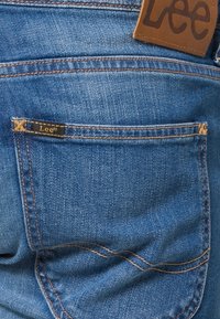 Blå denimjeans med en bakficka som har en guldig logotagg och kontrasterande sömmar längs sömmarna. Mjuk textur med lätt blekning.