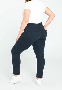 Jean skinny bleu foncé à taille haute, texture lisse, avec poches arrière. Associé à des baskets blanches pour un look décontracté.