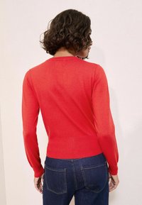 Kvinna med lockigt hår som vänder bort, iklädd en röd långärmad tröja och mörkblå jeans mot en ljus bakgrund.