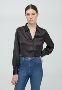 Camicetta cropped nera in satin con colletto, maniche lunghe e chiusura frontale a bottoni, abbinata a jeans in denim blu a vita alta.