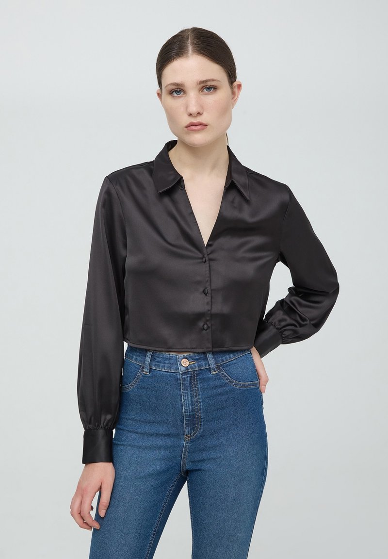 Camicetta cropped nera in satin con colletto, maniche lunghe e chiusura frontale a bottoni, abbinata a jeans in denim blu a vita alta.
