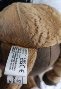 Jouet en peluche marron avec une fourrure courte et une texture douce. Il présente une étiquette cousue avec des informations sur le produit attachée à sa partie inférieure.