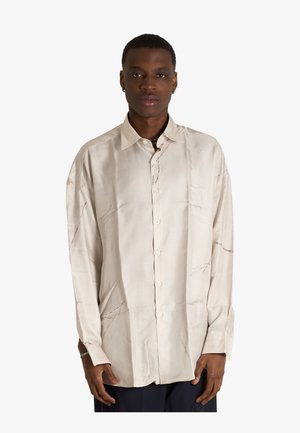 Giovane uomo che indossa una camicia beige larga con bottoni e pantaloni scuri, in piedi davanti a uno sfondo bianco.