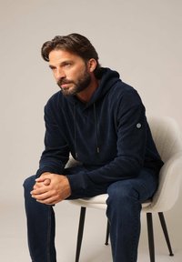 Haut à capuche en cordon bleu marine avec des lacets, associé à un jean bleu marine. Le tissu a une apparence texturée ; logo sur la manche. Assis sur une chaise.