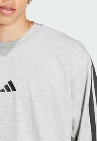 Grå sweatshirt med rund hals, der har et sort logo og stribede sorte detaljer på ærmerne. Blødt, let materiale.
