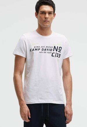 Man wearing white short-sleeve t-shirt with black and green text "game.set.match CAMP DAVID rule the court NO C|19" and dark pants, neutral background.