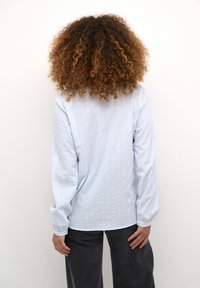 Ljusblå långärmad skjorta med vertikala vita ränder och textilstruktur. Baksidan har en rundad nederkant och knäppta manschetter. Tillsammans med mörka byxor.