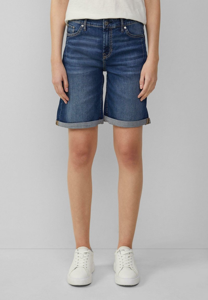 Denim Bermuda shorts in dark wash with rolled cuffs, five-pocket design, belt loops, and a button closure. Paired with white sneakers.
