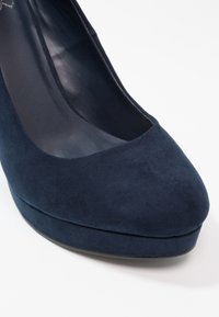 Chaussure à talon haut en daim bleu marine avec bout arrondi et semelle compensée, vue de face sur fond blanc.