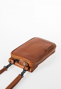 Braune Ledertasche mit querliegenden Riemen, mit Reißverschluss, flach-rechteckiger Form, schwarzen Hardware-Akzenten und einer seitlichen Schlaufe zur Befestigung.