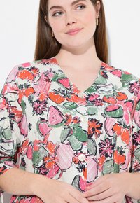 Blusa de seda con patrón floral en color crema con vibrantes tonos de rosa, verde y naranja. Presenta un escote en V y botones blancos. Textura de tela suave.
