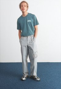 Téglazöld környakú póló "Dickies" logóval, szürke cargo nadrág zsebekkel, és szürke sportcipő fekete és sárga kiegészítőkkel.