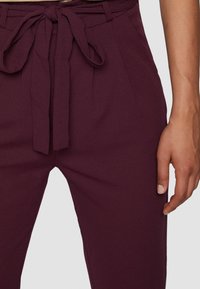 Burgunderfarbene Hosen mit einer taillierten Taille und einem Bindegürtel verfügen über eine glatte Stoffoberfläche und Falten, die ein maßgeschneidertes Design betonen.