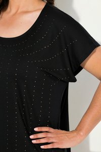 Černý top s krátkým rukávem vyrobený z měkké tkaniny, který se pyšní geometrickým vzorem malých růžově zlatých cvočků uspořádaných do vertikálních linií.
