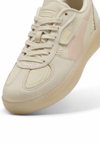 Beige színű sneaker texturált felsőrésszel, világos rózsaszín kiegészítőkkel az oldalon, valamint áttetsző talppal. Fűzővel és logóval rendelkezik a nyelvén.