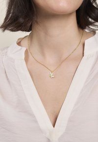 Collana in oro con pendente a forma di farfalla con una pietra bianca accentata, esposta su una blusa chiara con scollatura profonda.