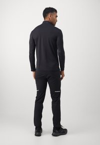 Black long-sleeve top with a high collar, paired with black pants featuring reflective accents and textured fabric. Worn by a male figure from behind.
