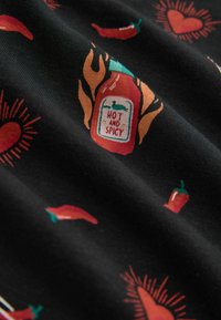 Schwarzer Stoff mit roten Chilischoten, Flasche mit feurig scharfer Sauce beschriftet mit „Hot and Spicy“ und Muster aus roten Herzen mit strahlenden Linien.