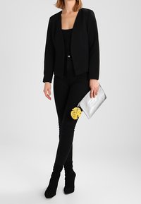 Blazer noir sur un haut noir, assorti à un jean skinny noir et des bottines à talons noires. Pochette argentée avec un détail accent en forme de feuille jaune.