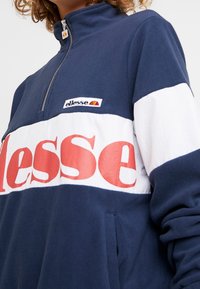Mörkblå fleece zip-up sweatshirt med ett stort vitt band med röd text "ellesse" och en liten logotyp på övre vänstra sidan.
