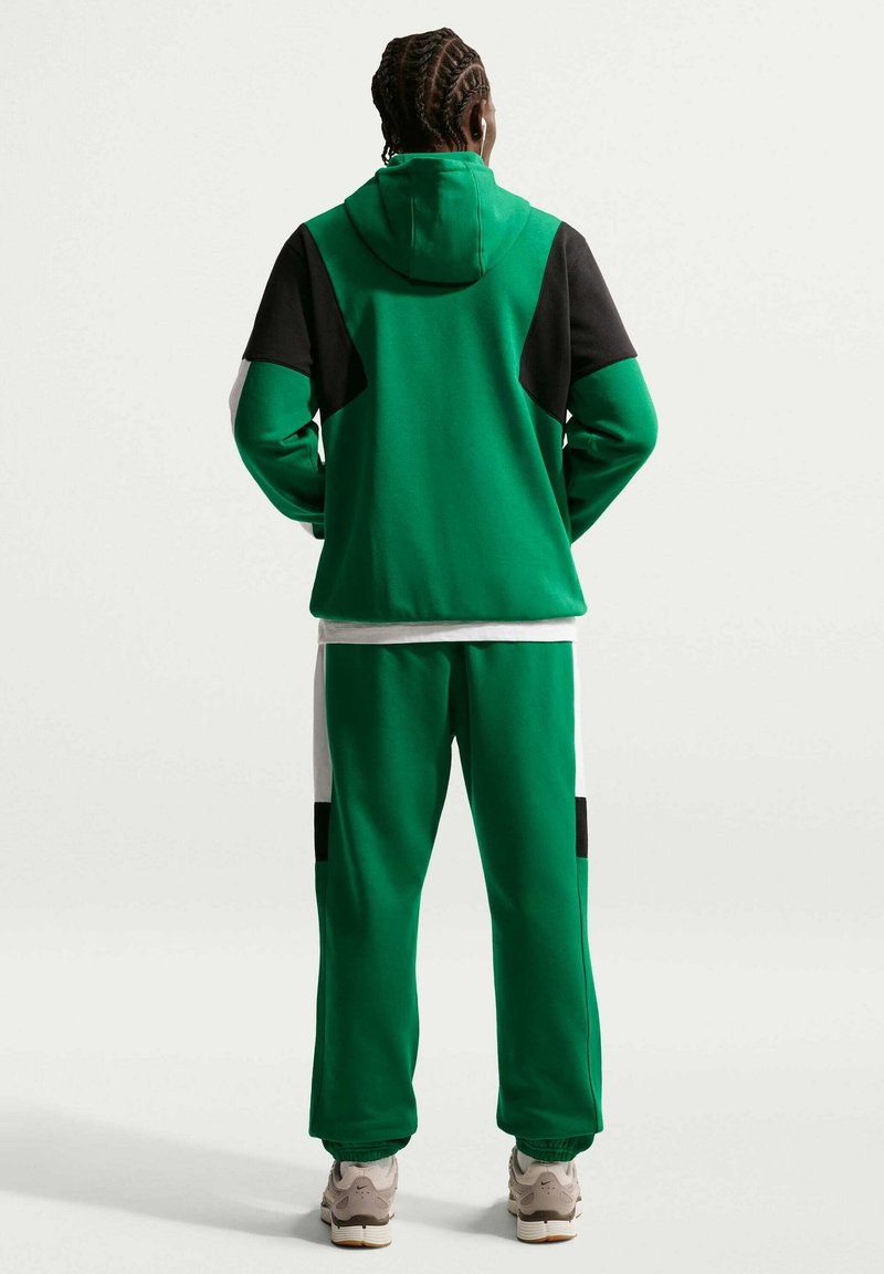 Persona in piedi di spalle alla telecamera che indossa una tuta verde con pannelli neri e bianchi e sneakers beige su sfondo uniforme.